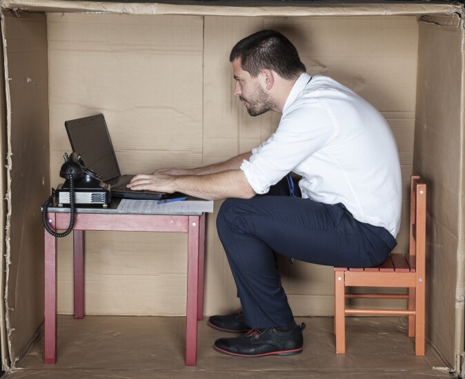 Businessman hunched over inside a small office made from a cardboard box.
