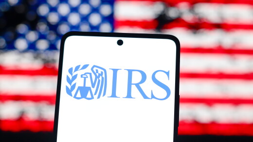 In this photo illustration, the IRS logo is displayed on a smartphone screen and in the background the flag of the United States