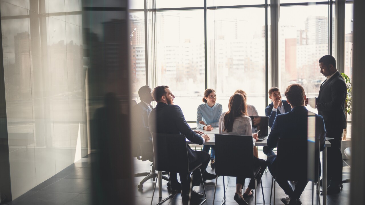 Eight professionals are sitting at a conference table, listening to a colleague speak.
