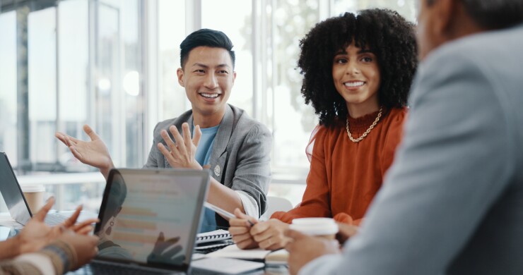 An Asian man and a Black woman listen excitedly to their colleague who is sitting at the head of a conference table; just his shoulder is in view.