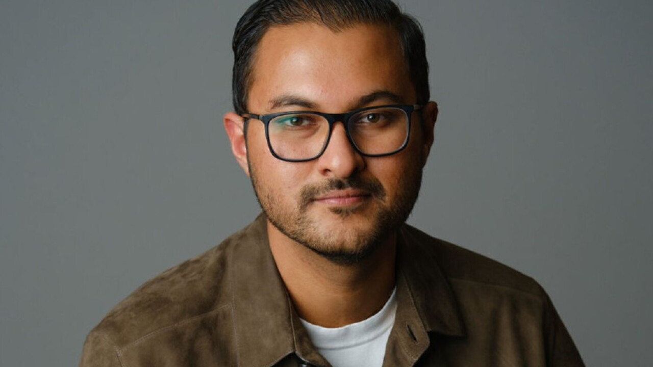 Headshot of a man with black glasses and short beard, wearing a brown suede shirt against a solid gray background.