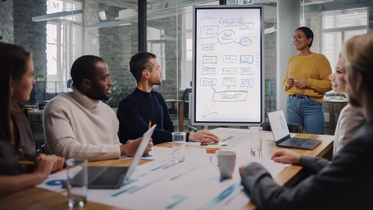 Project Manager Makes a Presentation for a Young Diverse Creative Team in Meeting Room in an Agency. Colleagues Sit Behind Conference Table and Discuss Business Development, User Interface and Design