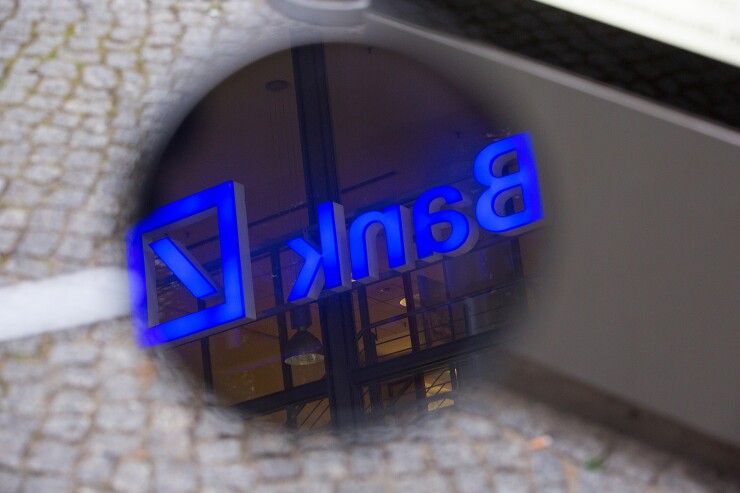 An illuminated logo for Deutsche Bank reflects in the mirror of a motor scooter as its sits parked outside a bank branch in Berlin.