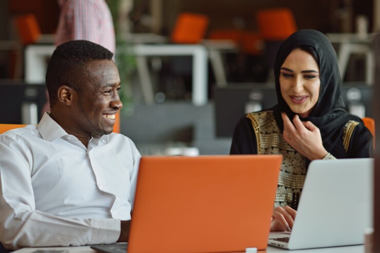 A Black man and Muslim woman are sitting at their desks, smiling.