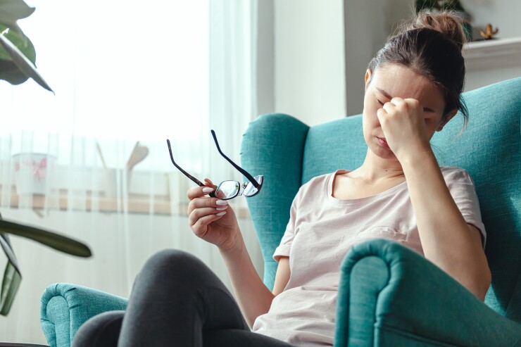A woman is sitting in a teal blue armchair, rubbing her temples in exhaustion with her glasses in her other hand.