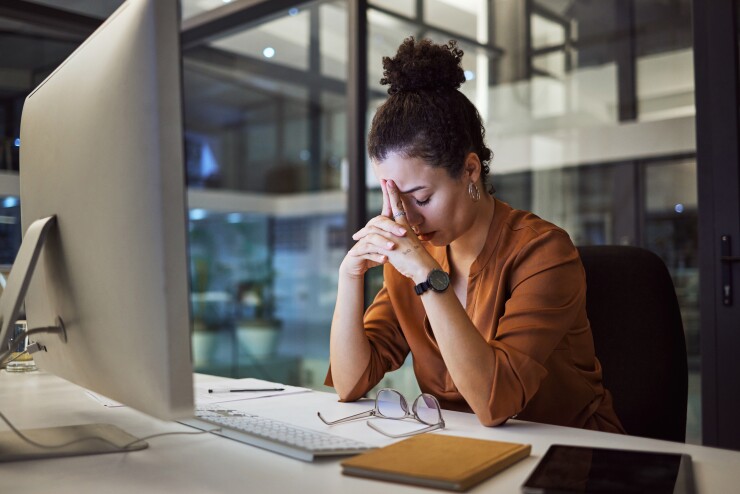 A young woman is at her desk in front of a large desktop computer; her head is down in exhaustion.