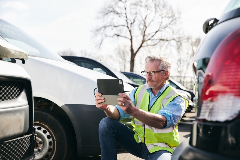 Loss adjuster wearing hi-vis safety vest standing in compound for damaged cars taking photo for insurance claim report on digital tablet