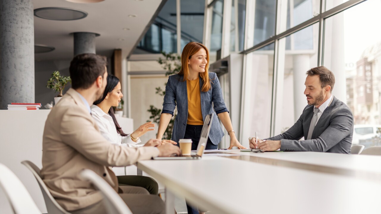 Woman standing at table, smiling, speaking to seated employees