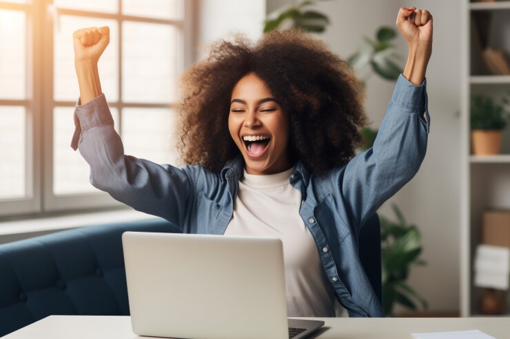 A woman puts her hands up in celebration in front of her laptop.