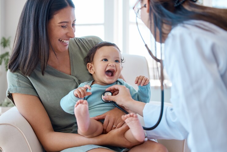 A mom is sitting with her baby while a doctor listens to the baby's heartbeat with a stethoscope.