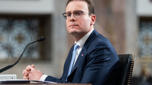 A man in a suit and glasses sits at a desk with a microphone, looking forward with a serious expression in a formal room.