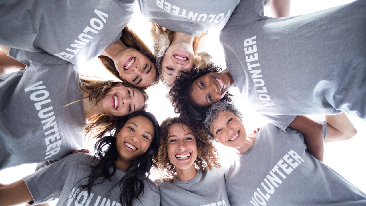 A group of women with grey t-shirts that read "volunteer" stand above a camera with their heads touching and their arms interlocked in a circle