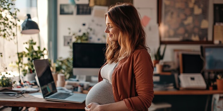 Pregnant woman, hands on stomach, standing near desk with computer, peaceful