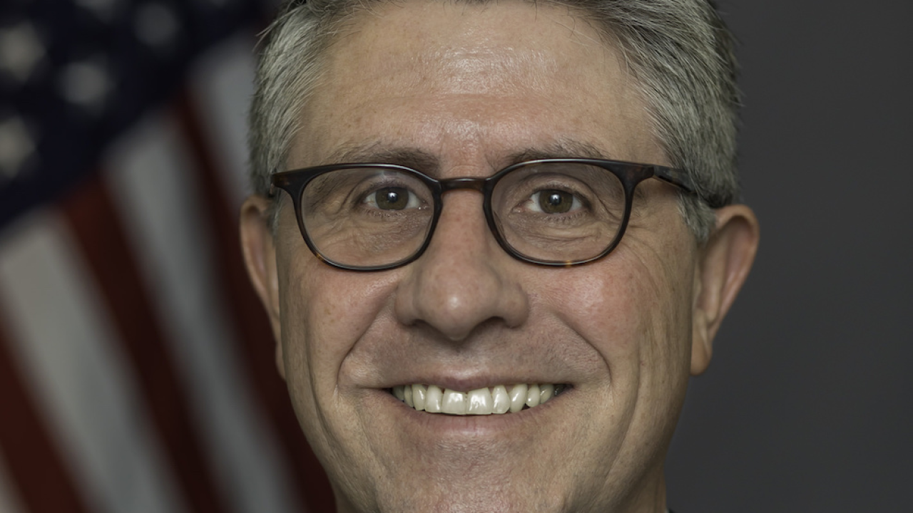 Headshot of man with gray hair and glasses wearing a suit and patterned pink tie standing in front of a blurred American flag in the background.