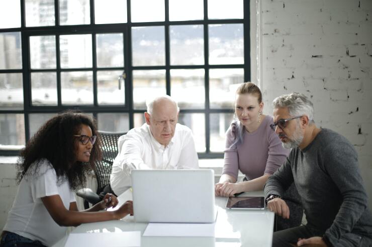Two young women and two older men sit in an office, all speaking at a laptop, presumably in a group meeting with their virtual colleagues.