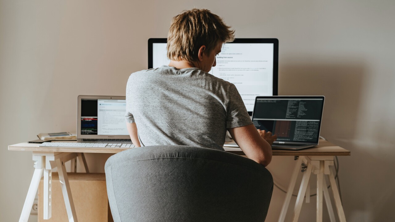 A man sits at a desk with three computer monitors. His back is to the reader, and it's implied he's working from home.