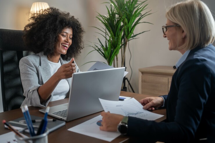 Two women sitting across from each other at desk, laughing, looking at computer and paperwork