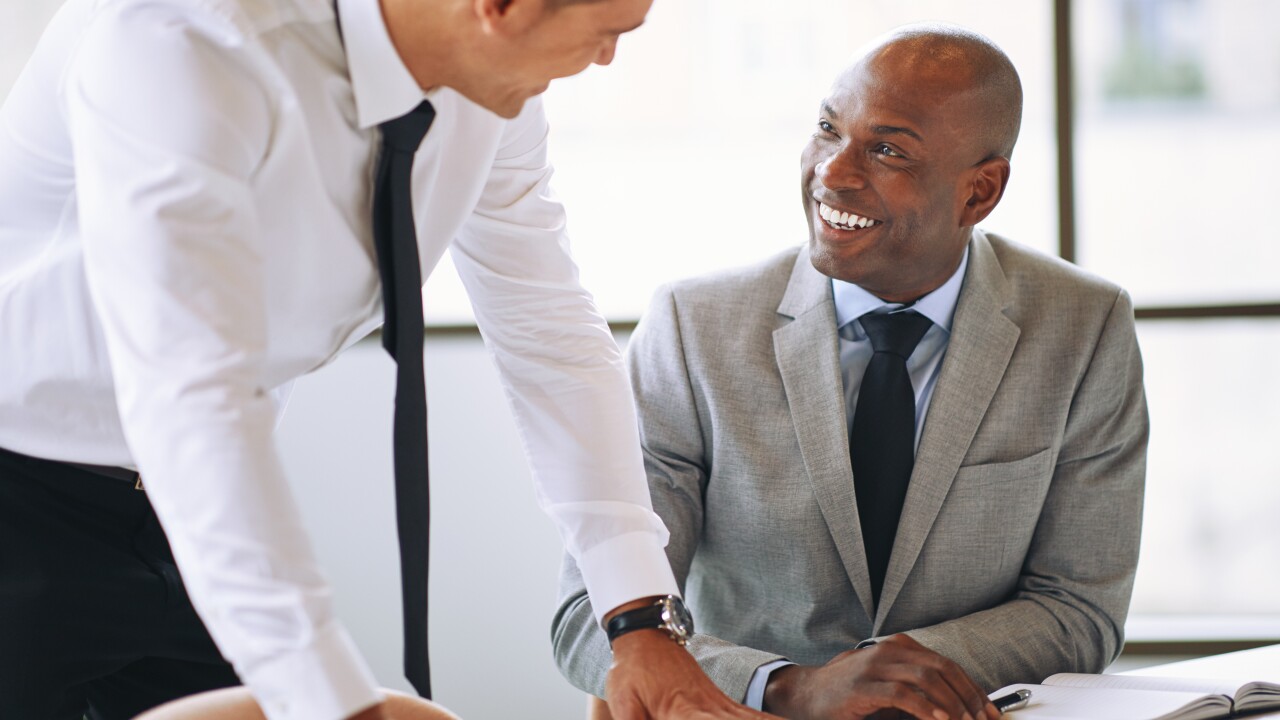 Two male employees talking at work, one seated, smiling