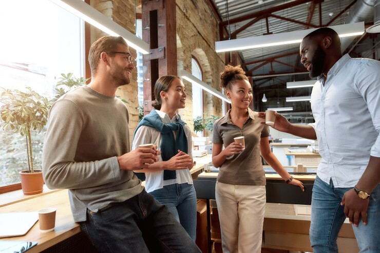 Four employees standing in office kitchen with coffee talking and laughing
