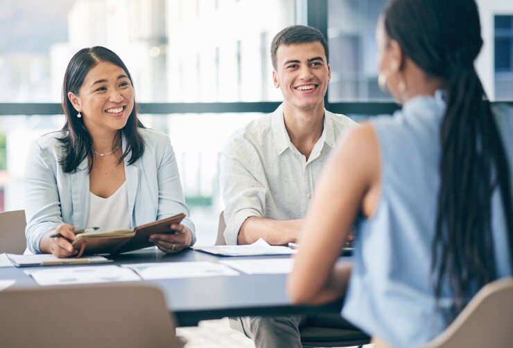 A young Asian woman and white man smile at a Black woman sitting across from them at a conference table.