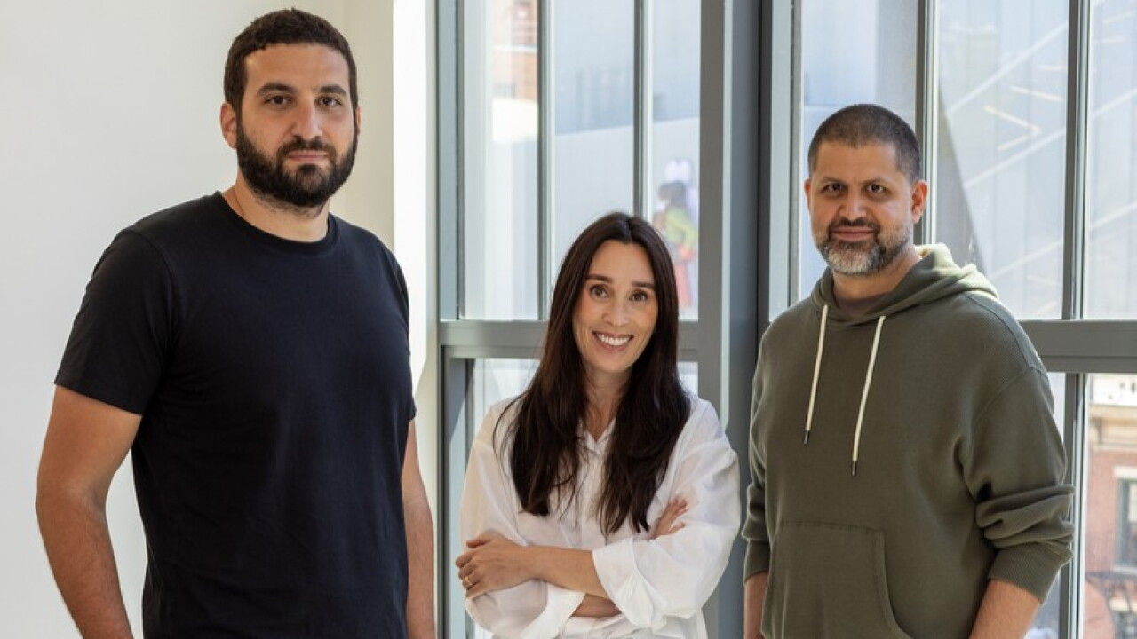 Three people standing by large windows: a smiling woman in white flanked by two men in a black t-shirt and green hoodie.