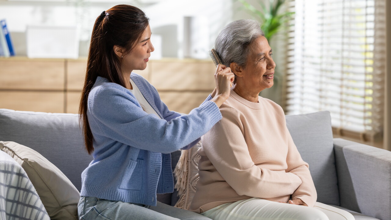 Young woman caring for elderly mother adjusting hearing aid device on couch at home, expressing warmth, love, compassion, and family connection in comfortable bright living room environment.