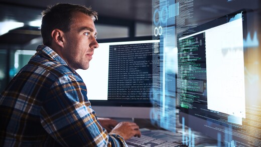 A person looking at two monitors with a holographic image of code.