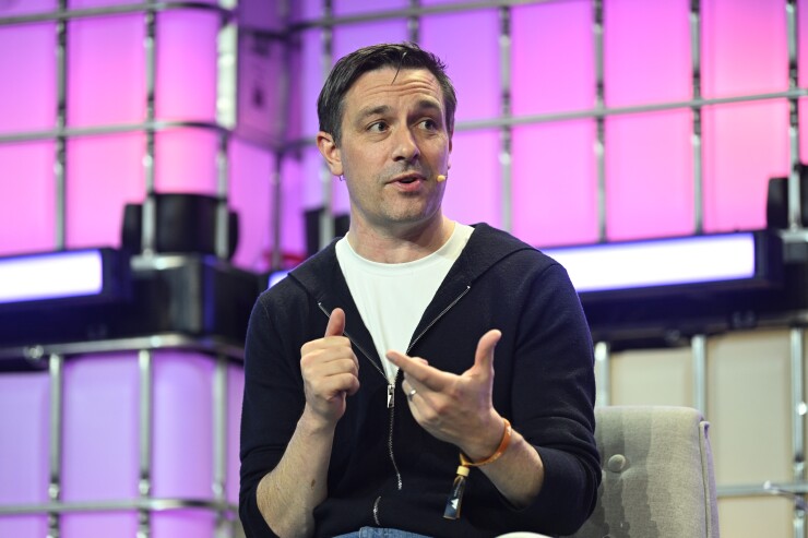 A man with short dark hair gestures while speaking on stage with a purple grid background, wearing a black zip-up hoodie over a white t-shirt.