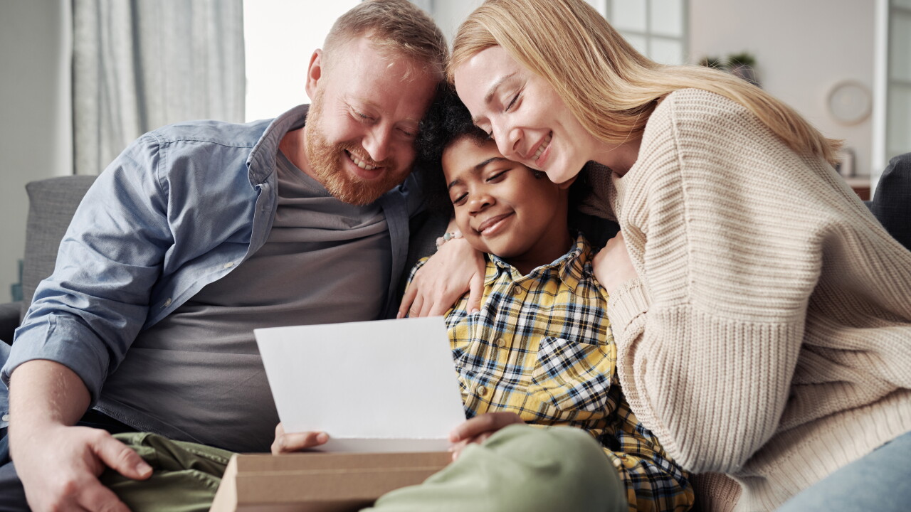 Happy parents embracing their African adoptive son while reading adoption agreement together on sofa.