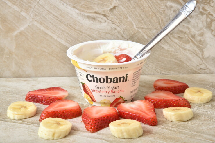 Chobani's strawberry banana yogurt sitting on a table surrounded by strawberry and banana slices.