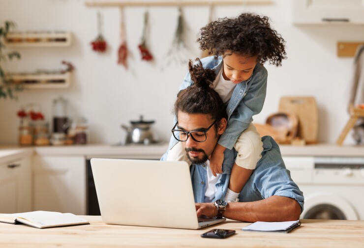 A Black dad with his son on his shoulders is working at his kitchen counter.