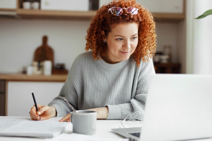 Portrait of happy female student watching webinar or tutorial studying online on laptop sitting at kitchen table, noting down after teacher or lecturer, looking at screen with interest